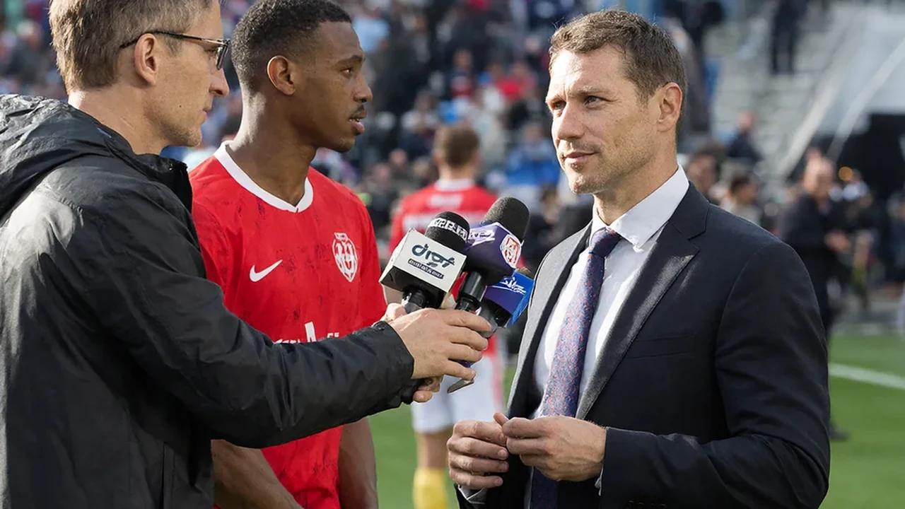 Manager speaking to reporters at a press conference while a player trains behind him